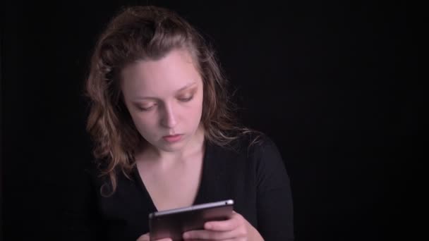 Retrato de la joven envuelta de pelo rizado mirando atentamente en la tableta y sonriendo con gusto en la cámara en el fondo negro . — Vídeo de stock