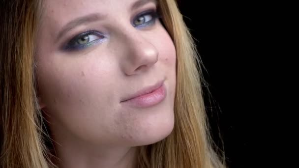 Zbliżenie strzelać z młody piękny żeński twarz z oszałamiający makijaż patrząc w camerawith wyrafinowany uśmiech w tle na czarno — Wideo stockowe