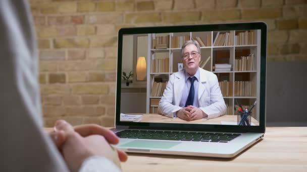 Online videocall van senior Doctor in witte Lab vacht en bril praten serieus aan zijn patiënt achter het scherm. — Stockvideo