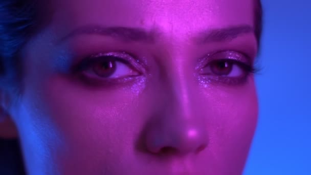 Close-up shoot of futuristic fashion model in colorful neon lights watching fixedly into camera in studio. — Stock Video
