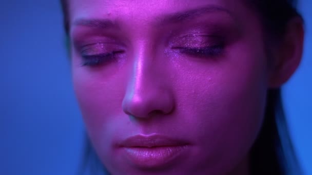 Futuristický model s třpytím make-upu v purpurově neonové světlech bliká a ve studiu pokojně sleduje kameru. — Stock video