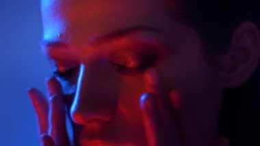 Yüzüne dokunan kırmızı ve mavi neon ışıklı modelin yakın çekim çekimi kameraya sabit olarak saatler.