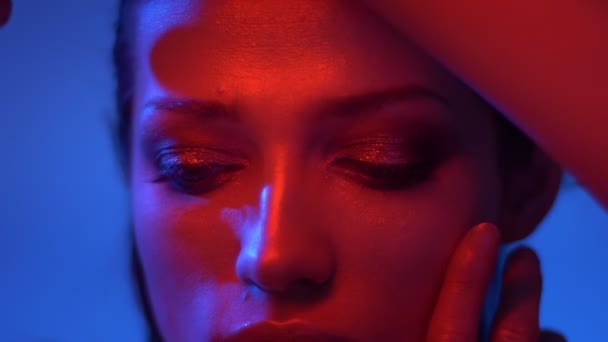 Close-up shoot of fashion model in red and blue neon lights hiding her face with fingers and watches into camera. — Stock Video