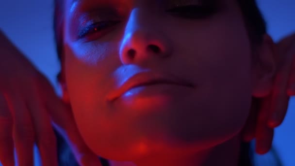 Zbliżenie modelu mody w czerwone i niebieskie neonowe światła dotykając jej twarzy i zegarki zapraszająco do aparatu. — Wideo stockowe