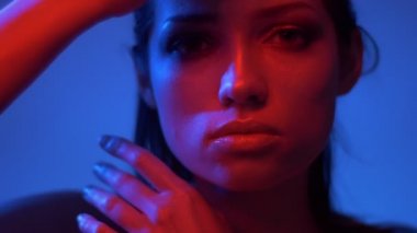 Kırmızı ve mavi neon ışıklarla fütüristik moda modeli eller ile yüze hafifçe dokunuyor ve kameraya sakince bakarak.