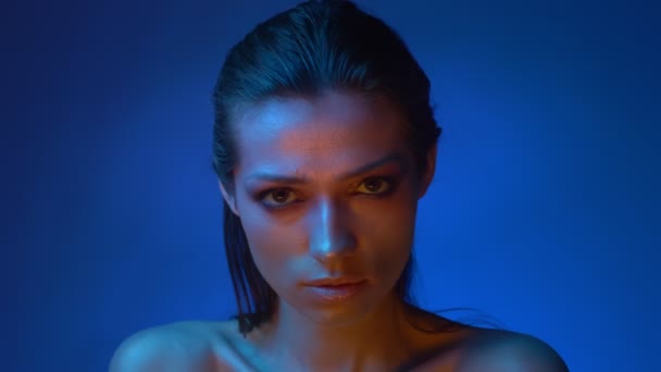 Сяюча модель з блискучим макіяжем, що стоїть у блакитних неонових вогнях, підозріло дивиться в камеру в студії . — стокове відео
