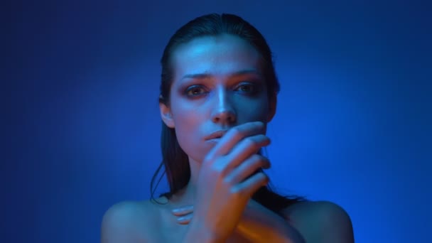 Modèle de mode futuriste brillant dans des néons bleu froid touchant et couvrant son visage avec les mains sur fond bleu . — Video