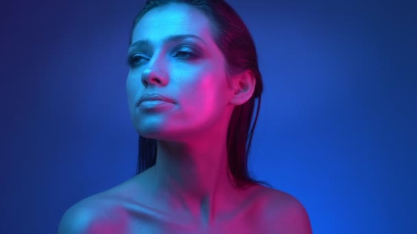 Glühendes Model mit glitzerndem Make-up in blauen und rosafarbenen Neonlichtern, das die Umgebung im Studio genau beobachtet. — Stockvideo