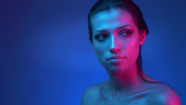 Świecący model z błyszczącym blasku makijażu w niebieskich i różowych lampach neonowych oglądając uważnie rightwards w studiu. — Wideo stockowe