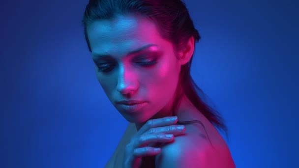 Świecący model z błyszczącym blasku makijażu w niebieskich i różowych lampach neonowych oglądając w aparacie dotyka jej ramię czule. — Wideo stockowe