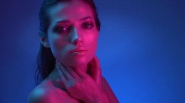 Mavi ve pembe neon ışıklarla ışıltılı parlak makyajlı profilde parlayan model, kameraya nazik bir şekilde poz vererek boynuna dokunuyor.