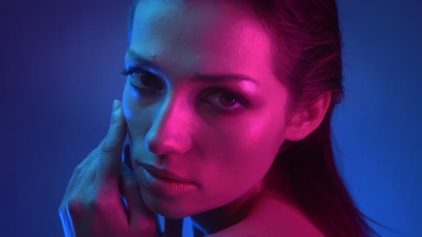 Close-up shoot of model in blue and pink neon lights watching over shoulder into camera with flirting glance and touching her face tenderly. — Stock Video