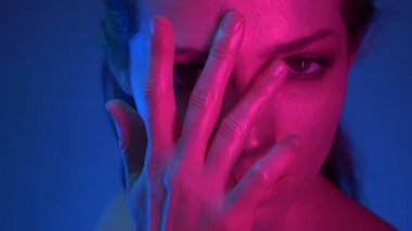Mavi ve pembe neon ışıklı ışıltılı makyajlı modelin yakın çekim çekimleri, parmaklarını kameraya hevesle izleyerek yüzünü kaplıyor.