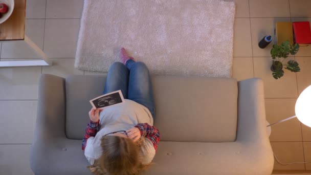 Hlavní střelka mladé těhotné ženy diskutuje o rentgenovém obrazu jejího dítěte, které mluví na telefonu, zatímco sedí na pohovce uvnitř — Stock video