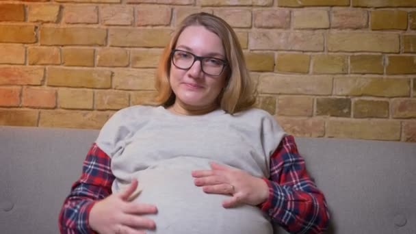 Closeup shoot of young pregnant female gently holding her stomach while sitting on the couch looking at camera and smiling in a cozy apartment indoors — Stock Video