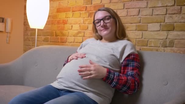 Primer plano de la joven embarazada sosteniendo suavemente su estómago mientras está sentada en el sofá mirando a la cámara y sonriendo felizmente en un acogedor apartamento en el interior — Vídeo de stock