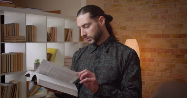 Sesión de primer plano de joven atractivo musulmán barbudo estudiante masculino leyendo un libro en una biblioteca mirando a la cámara interior — Vídeo de stock