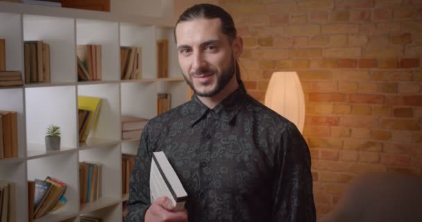 Closeup shoot of young attractive muslim bearded male student holding a book in a library looking at camera indoors — Stock Video