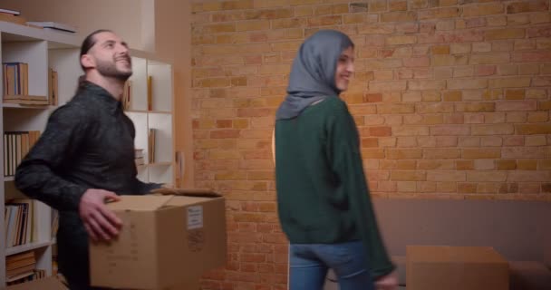 Primer plano de la joven pareja musulmana alegre caminando en un apartamento recién comprado e inspeccionando una habitación abrazando y sonriendo felizmente de pie junto a las cajas — Vídeo de stock