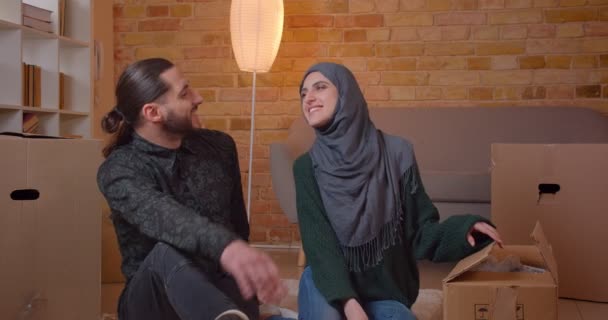 Primer plano de la joven pareja musulmana feliz sentada en el suelo junto a las cajas en un apartamento recién comprado sonriendo alegremente aconsejando dónde colgar una foto — Vídeos de Stock