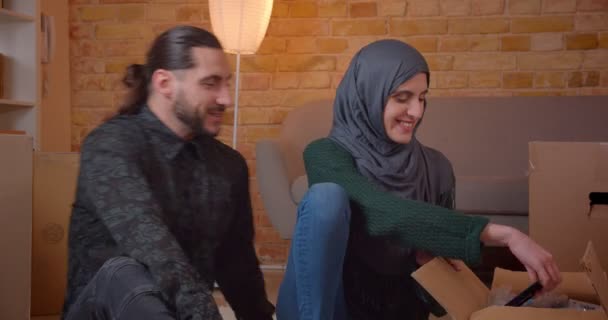 Tiro de close-up de jovem casal muçulmano alegre sentado no chão ao lado das caixas em um apartamento recém-comprado sorrindo feliz aconselhando sobre onde pendurar um quadro — Vídeo de Stock