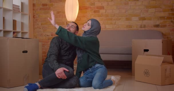 Closeup shoot of young cheerful muslim couple sitting on the floor next to the boxes in a newly bought apartment smiling happily dreaming about how to furniture the room — Stock Video