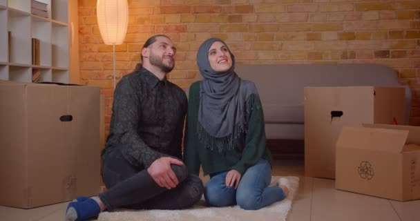 Primer plano de brote de joven pareja alegre musulmán sentado en el suelo junto a las cajas en un apartamento recién comprado soñando con cómo muebles de la habitación — Vídeo de stock