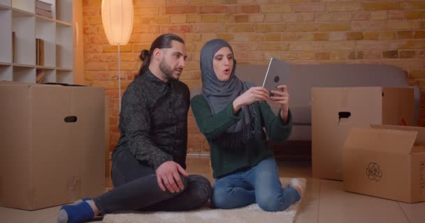 Neşeyle gülümseyen tablet kullanarak yeni satın alınan bir dairede katta oturan genç mutlu Müslüman çift Closeup ateş — Stok video