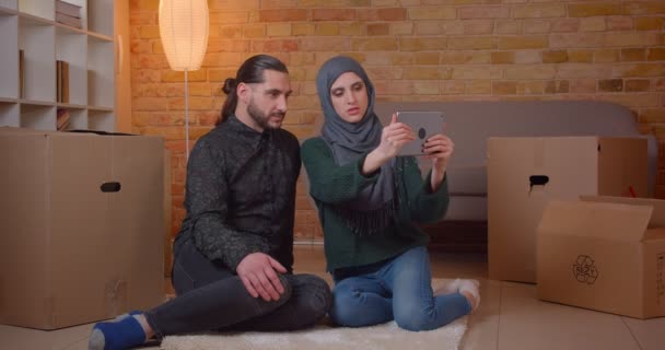 Yeni satın alınan bir dairede kutuların yanında yerde oturan genç mutlu Müslüman çiftin neşeyle gülümseyen tableti kullanarak yakın çekim — Stok video