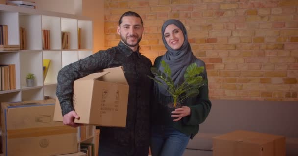 Retrato de primer plano de una joven pareja musulmana alegre mirando la cámara de pie en un apartamento recién comprado — Vídeo de stock