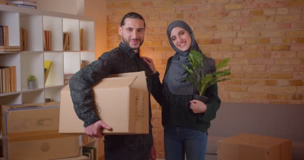 Retrato de cerca de una joven pareja musulmana alegre mirando la cámara de pie en un apartamento recién comprado sonriendo felizmente — Vídeo de stock