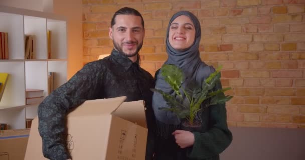 Retrato de cerca de una joven pareja alegre musulmana mirando a la cámara de pie en un apartamento recién comprado sonriendo felizmente. Hombre sosteniendo una caja y mujer sosteniendo un floerpot — Vídeo de stock