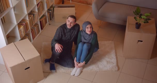 Крупный план съемки молодой счастливой мусульманской пары, сидящей на полу в недавно купленной квартире, весело улыбающейся и мечтающей о том, как обставить комнату в помещении — стоковое видео
