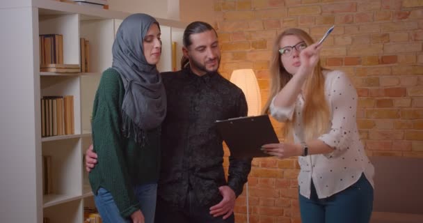 特写拍摄年轻的快乐穆斯林夫妇交谈，在一个新买的公寓拥抱欢快室内的房地产经纪人 — 图库视频影像