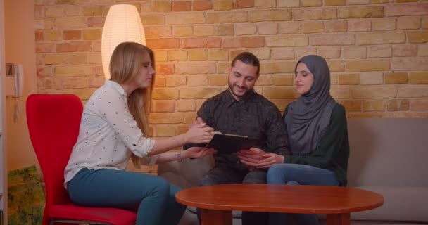 Zbliżenie strzelać z młody szczęśliwy muzułmański para omawiając z a realter a nowy Apartament uśmiechnięty wesel siedzący na kanapie w pomieszczeniu — Wideo stockowe
