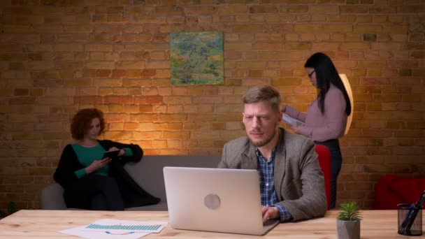 Крупним планом знімок дорослого бізнесмена, який друкує на ноутбуці, збуджується і святкує з двома жінками-колегами в приміщенні в офісі — стокове відео