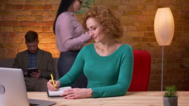 Dizüstü bilgisayarda yazan yetişkin iş kadınının kameraya bakarak not lar alması ve ofiste kapalı mekanlarda gülümsemesi. Arka planda yürüyen kadın ve erkek çalışanlar
