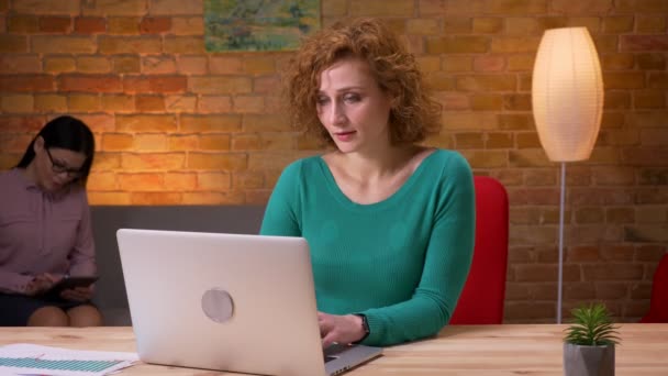Closeup shoot of adult businesswoman typing on the laptop looking at camera and smiling indoors in the office. Female employee using the tablet on the background — Stock Video