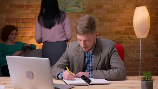 Nahaufnahme eines erwachsenen Geschäftsmannes, der im Büro mit dem Laptop Notizen macht. Mitarbeiterin sitzt mit Tablet auf der Couch und diskutiert mit einem Kollegen im Hintergrund — Stockvideo