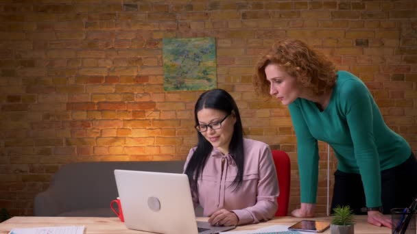 Primer plano de la sesión de dos mujeres de negocios discutiendo datosen el portátil en el interior de la oficina. Empresario pasando y saludando — Vídeos de Stock