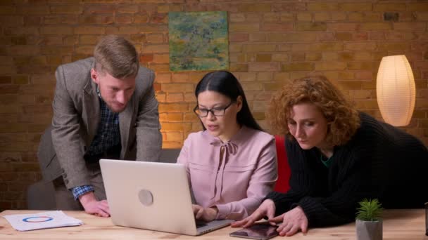 Tiro de close-up de jovem empresária asiática trabalhando no laptop celebrando o sucesso com dois colegas. Funcionário feminino segurando um tablet — Vídeo de Stock