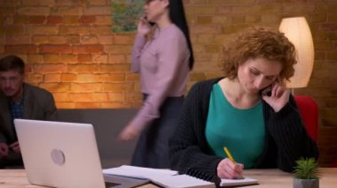 Yetişkin iş kadını, dizüstü bilgisayarda telefonla arama ve ofiste kapalı notlar alarak yazarak closeup ateş. Grafikleri ve arka planda yürüyen kadın çalışan ile veri getiren iş arkadaşı