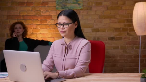 Zbliżenie strzelać z dorosły Asian businesswoman praca na The laptop mający a rozmowa telefoniczna i relaksujący wewnątrz w biurze. Kobieta pracownik dzwoniąc na telefon komórkowy na tle — Wideo stockowe