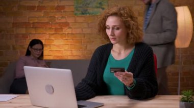 Kredi kartı ile dizüstü bilgisayarda online yetişkin iş kadını alışveriş closeup ateş ve ofiste mutlu kapalı gülümseyerek. Arka planda cep telefonu kullanan kadın çalışan