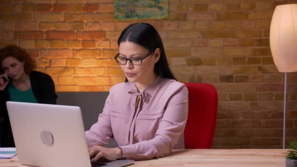 Close-up tiro de adulto asiático empresária trabalhando no laptop ter um telefonema e relaxante sorrindo alegremente dentro de casa no escritório. Funcionária telefonando para o celular em segundo plano — Vídeo de Stock