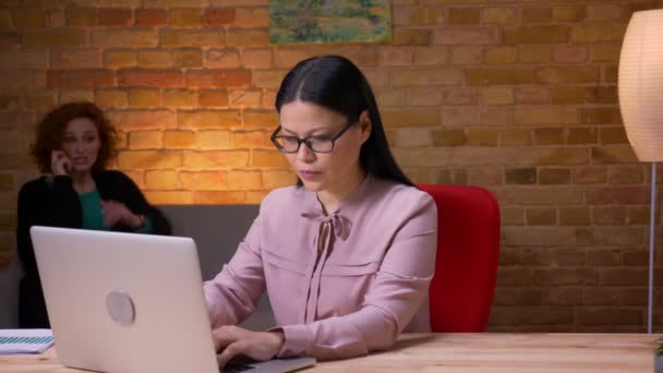 Zbliżenie strzelać z dorosły Asian businesswoman praca na The laptop mający a telefon rozmowa relaksujący patrząc w aparat fotograficzny i uśmiechnięty wnętrze w The biuro — Wideo stockowe