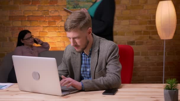 Close-up shoot van volwassen zakenman met behulp van de laptop ontvangen van een melding op de telefoon en glimlachend gelukkig binnen op kantoor. Vrouwelijke werknemer belt op de mobiele telefoon op de achtergrond — Stockvideo