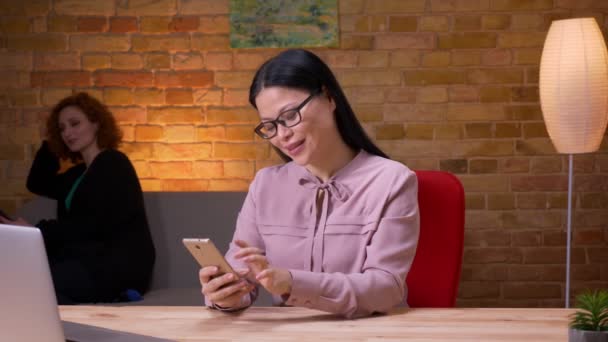 Съемки крупным планом взрослой азиатской предпринимательницы, делающей селфи по телефону в помещении офиса. Женщина-работница, использующая планшет на заднем плане — стоковое видео