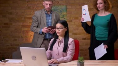 Ofiste dizüstü bilgisayar üzerinde çalışan yetişkin Asya iş kadını Closeup ateş. İşvereni ile görüntülü görüşme yapan üç iş arkadaşı, grafik vurgulandığı bir rapor hazırlıyor