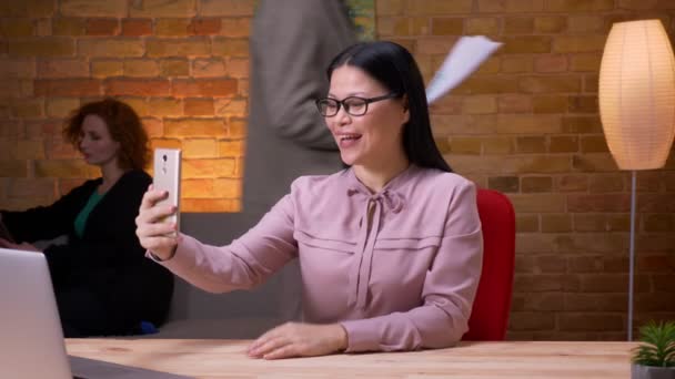 Närbild shoot av vuxna asiatiska affärskvinna med ett videosamtal på telefonen inomhus på kontoret. Kvinnlig anställd använder tabletten på bakgrunden — Stockvideo